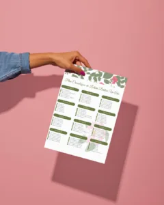 Mano sosteniendo plan cronológico de lectura bíblica en PDF con detalles de hojas en verde oliva sobre un fondo rosa.