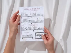 Manos sosteniendo hoja impresa con el tracker de libros de la Biblia en repisas para colorear sobre fondo de tela blanca.
